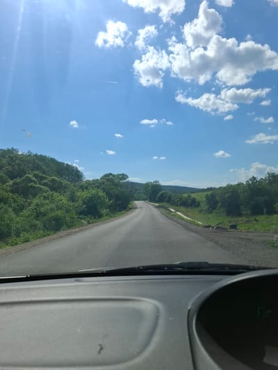 The image shows a view from inside a vehicle, possibly a car, driving on a paved road. The road is surrounded by lush green trees and vegetation on both sides. In the distance, there is a hill or mountain. The sky is blue with scattered white clouds. The vehicle's interior is visible on the right side of the frame. No people are visible. The location is likely a rural area near Baylovo, Bulgaria, given the landscape.