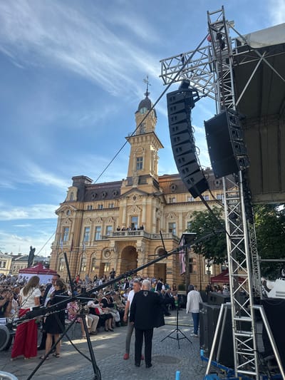 The image depicts an outdoor event taking place in front of a large, ornate yellow building with a clock tower, identified as the building of the Municipal Office in Nowy Sącz, Poland. A large outdoor stage structure with speakers and lighting equipment is set up on the right side of the frame, partially obscuring the view of the building. A crowd of people are seated and standing in front of the stage and around the plaza. Several individuals are standing near the stage setup, some interacting with each other, while others are seated in the audience. The sky is blue with wispy clouds. The overall scene suggests a public gathering or festival.