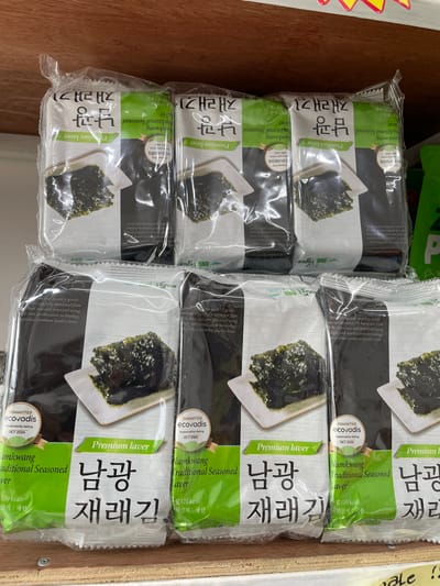 The image shows a shelf in a store, possibly a supermarket or a convenience store, displaying several packages of dried seaweed snacks. The products are stacked in two rows. The top row displays three packages of seaweed labeled "Premium laver" with Korean text. Below these, a second row shows more packages of the same product, also labeled "Premium laver" and "Namkwang Traditional Seasoned Laver." The packaging is predominantly green and white, with images of the seaweed snacks. The items are neatly arranged on what appears to be a wooden shelf. The lighting suggests an indoor retail environment. The overall scene is a typical retail display of food products.
