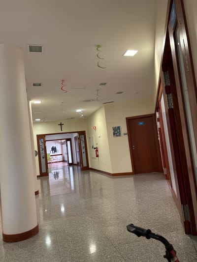 This image depicts an indoor hallway or corridor within a building in Santa Maria da Feira, Portugal. The floor is covered with light gray, speckled terrazzo tiles, which are highly reflective, showing ambient light sources and reflections of distant objects. The walls are painted a light cream color, transitioning to a white ceiling.

Numerous square and rectangular recessed light fixtures illuminate the ceiling, from which red and green spiral streamers are suspended at various intervals along the hallway. A large, round, off-white pillar with a dark brown wooden base trim stands prominently on the left side of the frame.

At the far end of the hallway, a black crucifix is mounted on the wall above a set of double wooden doors featuring glass panels. Through these doors, a brightly lit room is visible, containing a dark, two-dimensional silhouette cutout depicting two seated figures on a bench. Reflections of these figures are evident on the polished floor surface.

Along the right-hand wall, several dark brown wooden doors are present. One of these doors displays a small blue sign. To the right of the double doors at the end of the hall, a red fire extinguisher is mounted on the wall, positioned beneath a red fire safety sign. A framed picture, possibly with a floral design, hangs on the wall between two doors on the right. The partial view of an open dark wooden door frame is visible on the extreme right of the image. The black handlebars of a bicycle or scooter are partially visible in the bottom right foreground. No individuals are immediately present in the visible foreground or midground of the hallway.