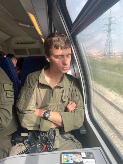 The image shows a young man wearing a green military uniform sitting on a train in a window seat. He is asleep with his eyes closed and arms crossed in front of him. He is wearing a black wristwatch, a black earbud in his right ear, and a black belt with a firearm attached to it. The man's right arm is resting on a small table. On the table, there is a colorful phone case with a white sticker that says "Better late than never" in black lettering. Through the window, a set of railroad tracks can be seen outside the train. The tracks are next to a green grassy field. There are power lines and a large steel structure in the distance. This scene is likely taking place on a train traveling through a rural area of Israel. The latitude and longitude coordinates of the location are 31.985, 34.871, which are in the town of Ahi'ezer, Israel.