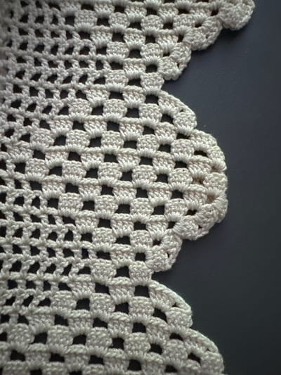 The image shows a close-up, high-angle shot of a crocheted doily with a scalloped edge. The doily is a pale cream color and is laid out on a dark gray surface. The crochet pattern consists of a repeating series of open squares and solid, clustered stitches that form a textured, floral-like design along the scalloped edge. The doily is positioned on the left side of the frame, with the dark gray surface occupying the right side. There are no people or discernible elements of a city or specific location present in the image.