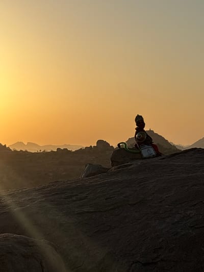 A medium shot shows a rock climber perched on a rock face at Kamalapura, India. The climber, facing the viewer, is wearing a red shirt, and climbing gear, including a helmet, climbing rope, and harness. The climber appears to be preparing for the next move. The rock face is a large, dark stone structure, partially obscured by shadows. The background features a light yellow and orange-hued sky, indicating sunset or sunrise. Rolling hills and other rock formations are faintly visible in the distance.