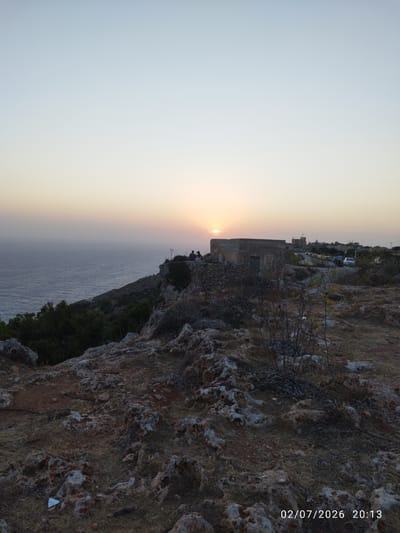 The image depicts a sunset view from Dingli Cliffs, Malta. The sun is low on the horizon, casting a warm glow over the sea. A few people are visible on the clifftop, enjoying the view. In the foreground, the rocky terrain of Dingli Cliffs is evident, with sparse vegetation. In the midground, there is an old stone building, and further in the distance, a road with some cars and another building can be seen. The sky is a pale blue with a hint of orange near the horizon. The timestamp "02/07/2026 20:13" is present in the lower right corner of the image.