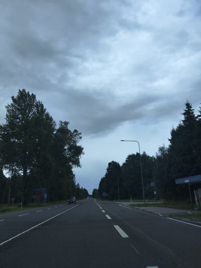 The image shows a road in Võiste alevik, Estonia, on an overcast day. The road is paved and has white lane markings. There are trees on either side of the road, and a few cars are visible. A blue road sign is visible on the left side of the road, and a bus stop shelter is on the right. The sky is cloudy and gray.