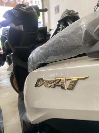 A white scooter with gold "Beat" lettering is parked indoors. The scooter is mostly in focus, with a plastic cover over part of the handlebars. Other scooters are visible in the background, along with Suzuki logos on the wall, suggesting the location is a dealership. The lighting is bright, indicating it might be daytime. The environment appears to be a showroom or garage.