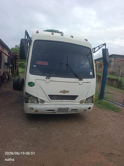 A white Chevrolet bus is parked on a gravel lot. The bus has a black grille, rectangular headlights, and a gold Chevrolet emblem on the front. There is a green sticker with the number "08" on the driver's side fender. The bus has a license plate that reads "03AABLD". In the background, there is a chain-link fence with blue and yellow poles. To the left of the bus, there is a building with a red roof and some tires. The sky is cloudy. The timestamp on the image indicates it was taken on June 19, 2026, at 06:51. The name "natha" is also visible on the image. The location is identified as El Chaparral, Venezuela.