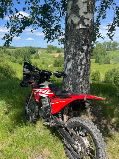 A red and white off-road motorcycle is parked, leaning against the trunk of a large birch tree on a sunny day in Новочувашский, Russia. The motorcycle, identified by "JHLR" branding on its handlebars and a "JHC" logo on its tank shroud, displays mud on its knobby tires and frame, indicating recent use. A grey and black motocross helmet rests on the handlebars. The prominent birch tree, positioned centrally, exhibits characteristic white bark with dark fissures and shadowed areas. Its green leaves from overhead branches frame the upper view of a blue sky with scattered white cumulus clouds. The immediate foreground consists of tall, verdant green grass interspersed with areas of sun and shadow. The midground features rolling green hills covered in dense deciduous trees and open grassy patches, through which multiple dirt tracks or trails are visible winding into the distance. The far background presents a hazy outline of what appear to be industrial structures or towers on the left horizon, beyond more distant tree lines. No individuals are present in the scene.