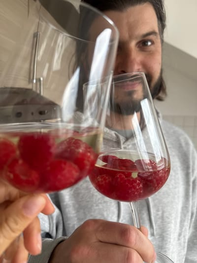 A close-up, low-angle shot shows a man holding two stemmed glasses filled with raspberries. The man has dark hair and a beard, and he is wearing a gray sweatshirt. His face is partially visible behind the glasses. The glasses are clear, and the raspberries are bright red. The background appears to be a kitchen with cabinet handles visible, and the lighting is bright. The location is unknown and there is no evidence of a location in the city of Ampfing, Germany.