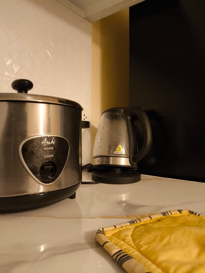 This is a close-up, eye-level shot of a kitchen counter in Imus, Philippines. The scene features a metallic rice cooker with a black lid and a black control panel, displaying the brand "Asahi" along with "Warm" and "Cook" buttons. To the right of the rice cooker is a clear glass electric kettle with condensation on its surface, indicating it has recently held hot water. A yellow and black striped pot holder rests on the counter in the foreground, partially obscuring the marble-patterned countertop, which shows some minor stains.

In the background, a white tile backsplash with a herringbone pattern is visible above the rice cooker. To the right of the backsplash, a wall transitions from a warm beige to a dark textured surface. A power outlet is visible behind the rice cooker. The lighting suggests it is likely daytime, possibly indoors with artificial lighting contributing to the scene's ambiance. There is no apparent activity taking place, the scene appears to be set up for use. The image conveys a sense of domesticity and everyday kitchen appliances.