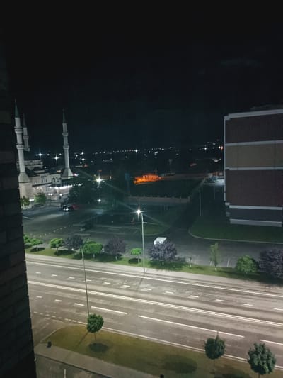 A night view of the Akhmad Kadyrov Mosque in Kurchaloy, Russia, is depicted from an elevated perspective. The mosque, characterized by its white minarets and light-colored building, is illuminated, creating a prominent focal point. Adjacent to the mosque is a multi-story building with brown and tan facades. The scene also includes a street with white lane markings, illuminated by streetlights. Several trees, some with dark foliage, are scattered throughout the area, lining the street and surrounding the mosque. The overall atmosphere is urban and tranquil, with the nighttime setting casting a soft glow on the illuminated structures and the surrounding landscape.