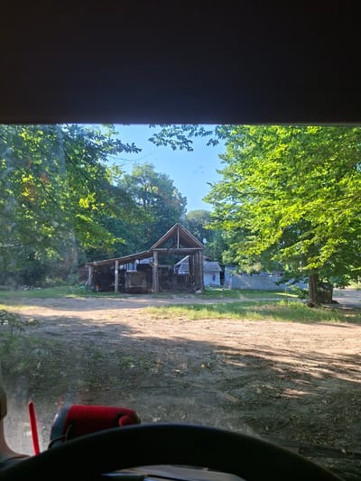 The image shows a view from inside a vehicle, looking out at a rustic wooden structure. The structure appears to be a shed or a small barn, with a pitched roof and open sides, revealing some items stored within. The building is situated in a rural setting, surrounded by lush green trees. A dirt road runs alongside the structure, with tire tracks visible on its surface, suggesting recent vehicle activity. Sunlight filters through the leaves of the trees, casting dappled shadows on the ground. The sky is a clear blue. On the right side of the frame, a part of the vehicle's interior is visible, including the steering wheel and a red object, possibly a seat or a piece of equipment. The scene is set during the daytime, with bright sunlight indicating fair weather. The overall impression is one of a peaceful, natural environment with a touch of rustic charm. The image was taken in Suruceni, Moldova.
