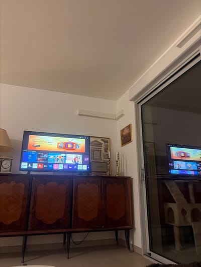 A modern living room presents a juxtaposition of classic and contemporary elements. An antique wooden cabinet with decorative panels stands beside a wall-mounted television. Above them, framed pictures adorn the wall below a large window reflecting the interior. A lamp and clock are visible near the cabinet's base.