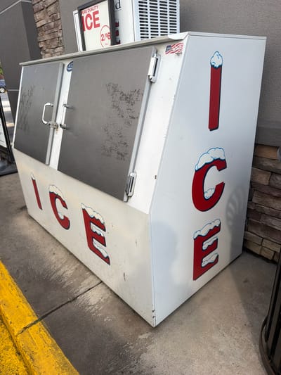 The image shows a large, white ice chest on a concrete surface in Raphine, United States. The ice chest has red lettering that spells "ICE" on both the side and the front. The lettering on the front is larger and more prominent. The lid of the ice chest is gray and has two handles. A sign with the words "PURE QUALITY ICE" and "2/ $6" is visible at the top of the ice chest. A "Made in USA" sticker is also present. The background shows a brick wall and a portion of a building with a gray facade. A yellow curb is visible in the foreground. The overall scene suggests an outdoor vending area for ice.