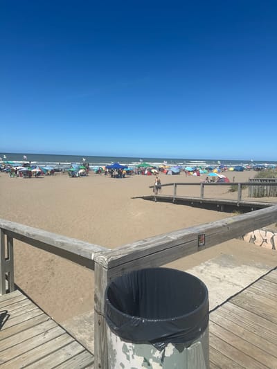 A view of a beach under a bright blue sky is partially obstructed by a wooden boardwalk and a black trash can liner. Numerous beach umbrellas dot the sand along the water's edge. The boardwalk runs parallel to the beach, with wooden planks and railings visible. The trash can liner is positioned on the boardwalk, close to the viewer.