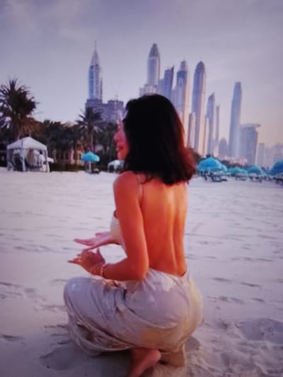 A woman with dark hair is seen from behind, kneeling on a sandy beach. She is wearing a light-colored, strapless top that exposes her back, and loose, flowing pants. Her hands are raised in front of her, with her palms facing outwards. In the background, several tall, modern buildings with pointed tops dominate the skyline, suggesting a city setting. Palm trees are visible among the buildings. A row of blue umbrellas and lounge chairs are set up on the beach, and white tents or cabanas are also present. The sky is a soft, hazy purple, indicative of dusk or dawn. The image does not depict the city of Prisel'e, Russia.
