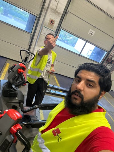 In a warehouse setting in Schweitenkirchen, Germany, two men are visible. The man in the foreground, with dark hair and a beard, wears a red shirt and a yellow safety vest with reflective stripes. He is looking directly at the camera. Behind him, another man in a white t-shirt and a yellow safety vest is holding a mobile phone in his left hand and making an offensive gesture with his right hand towards the camera. He is standing near a pallet jack. The environment appears to be a loading bay with large, overhead garage doors, one of which is marked with the number "10." A safety sign is visible on the wall. The floor is marked with yellow lines.