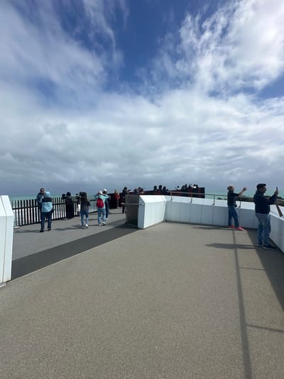 People stand along a modern boardwalk, some with arms raised, facing a distant view over a body of water. The structure is defined by clean, white geometric shapes and metal railings. A cloudy sky dominates the upper portion of the image. The scene conveys a sense of public gathering, perhaps a viewing point or promenade, with individuals looking out at a scenic landscape.