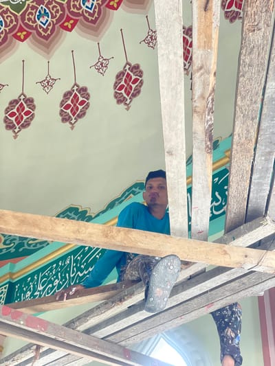 The image depicts the interior of a mosque or Islamic religious building under renovation or decoration in Mns Asan, Indonesia. A man is positioned on a multi-tiered wooden scaffolding structure, with wooden planks partially obscuring his body. He is wearing a blue long-sleeved shirt, heavily paint-stained camouflage-patterned trousers, and dark flip-flops. His right hand and forearm are stained with paint. He is looking directly at the camera with a puckered-lips expression. His right foot, with the sole of a dark flip-flop visible, is extended towards the viewer. Partially visible below and to the right are the lower legs of a second individual, also in paint-stained patterned trousers, indicating another worker on the scaffolding. The background features a light green ceiling extensively decorated with repeating Islamic geometric and stylized floral motifs in red, white, and brown tones. Below the ceiling, a wide band of dark green wall is adorned with prominent white Arabic calligraphy. The scaffolding consists of rough-hewn wooden planks and posts.