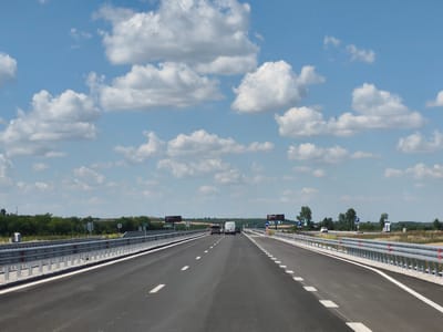 This image depicts a highway scene in Peshterna, Bulgaria, under a partly cloudy sky. The perspective is from the driver's seat, looking forward down a multi-lane asphalt road. The road is well-maintained with clear white lane markings. On either side of the road, there are metal guardrails with white posts and red reflectors. Beyond the guardrails, the landscape consists of flat terrain with sparse vegetation and a line of trees in the distance.
Several vehicles are visible on the highway. In the foreground, the road is empty. Further ahead, a white van is driving in the right lane, followed by a car. Another car is seen in the left lane. In the distance, there are highway signs, some of which are partially obscured. One sign displays the number "24". Other signs indicate parking areas with blue "P" symbols. A white portable toilet is visible on the right side of the road near the guardrail. The time of day appears to be midday, given the bright sunlight and the shadows cast. The weather is warm and pleasant, with scattered white clouds against a blue sky. There are no people visible in the image, only vehicles. The overall impression is that of a typical drive on a highway on a clear, sunny day.
