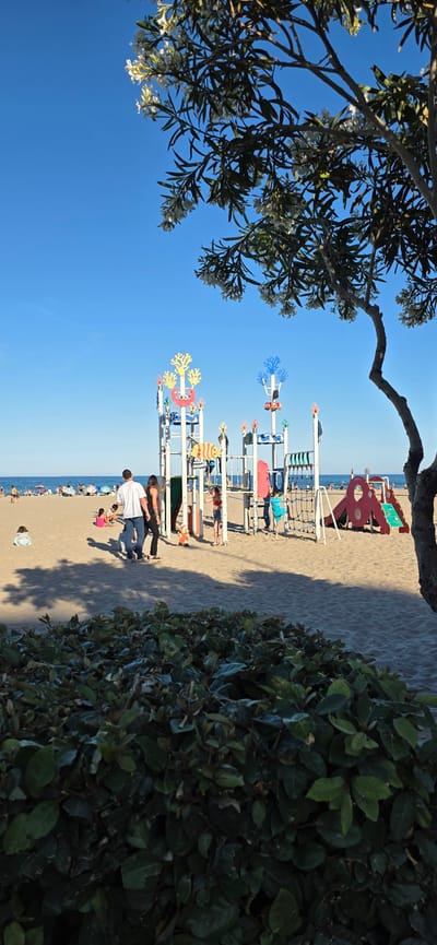 The image depicts a beach scene in El Port De Sagunt, Spain, on a sunny day. On the beach there is a playground, and people enjoying the beach. A couple with a child stand on the beach near the playground. Trees or bushes frame the view on the left and right sides.
