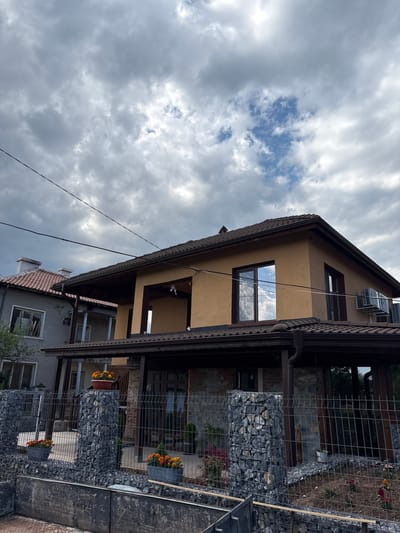 The image captures a residential street scene in Gorna Grashtitsa, Bulgaria, under an overcast sky with visible patches of blue. The primary subject is a two-story house with warm tan-colored upper walls and a dark brown tiled roof featuring an extended overhang supported by dark wooden beams. The lower facade of this house appears to be clad in a gray stone or textured material. On the upper floor, a large window on the left reflects the cloudy sky, while other windows are dark-paned. Two outdoor air conditioning units are mounted on the upper right exterior wall. A recessed balcony or porch with a dark ceiling is visible on the upper level, center-left.

To the immediate left, an older, two-story gray house with a reddish-brown tiled roof and a white chimney is partially visible. One upper-floor window of this adjacent house is open or damaged, revealing internal contents. A small, columned balcony extends from its front.

In the foreground, a gabion-style fence constructed from wire mesh cages filled with dark gray stones defines the property boundary. The fence includes gabion pillars supporting the mesh panels. Several dark-colored pots containing vibrant orange-flowering plants are placed atop the gabion structures and on the ground behind the fence. A dark, weathered concrete or metal structure runs horizontally across the very bottom of the frame, hinting at foreground construction or a low barrier.

Multiple dark power lines traverse the upper portion of the image against the sky. No individuals are present, and no discernible actions are occurring within the depicted scene.