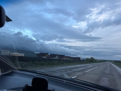 The image shows a view from inside a vehicle, looking out onto a road. The road appears to be wet and stretches into the distance, with a white dividing line visible. Beyond the road, there is a strip of green vegetation. Further on, there is a row of houses or buildings with pitched roofs. The sky is overcast with gray and white clouds, suggesting a cloudy day. The scene appears to be located near Столбова, Russia.