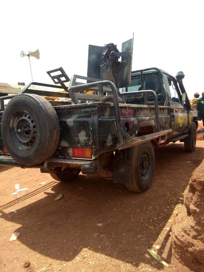 In Gana Daji, Nigeria, a dark green, camouflage-painted Toyota Land Cruiser pickup truck, modified for military or security operations, is parked on reddish-brown dirt. The vehicle is depicted from a low-angle, rear-three-quarter view, facing towards its front-right. A heavy machine gun with an armored shield is mounted in the truck bed, which is enclosed by a custom-fabricated metal roll cage and protective frame. A spare tire is secured vertically on the rear tailgate. A horn-shaped loudspeaker on a pole is affixed to the left side of the roll cage. The truck features a snorkel on its right side and a yellow circular decal on the passenger door, displaying a partially visible "GDB" or similar marking. The ground is composed of reddish-brown dirt with scattered small debris. In the background to the right, three individuals are partially visible; one wears a yellow garment, another a dark green garment, and they are standing near multiple orange containers. The sky above is bright and clear.