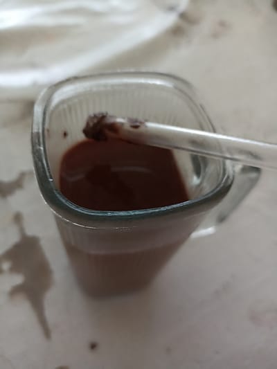 A close-up, overhead shot shows a clear glass mug filled with a dark brown liquid, likely hot chocolate or coffee, on a light-colored, speckled surface. A clear plastic straw rests across the rim of the mug, partially submerged in the liquid. The straw appears to have some of the brown substance clinging to its end. The surface around the mug is marked with several dark, irregular splatters. The background is out of focus, but suggests a textured, light-colored fabric or surface. The scene appears to be a casual setting, potentially a home or cafe, in Кош-Дөбө, Kyrgyzstan.