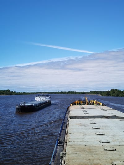 A daytime, medium-shot depicts a river scene near the city of Chaika, Russia. Two barges are visible on the water. One barge is positioned to the left, moving away from the viewer. The other is in the foreground. The latter occupies the bottom half of the frame, showing a gray deck with evenly spaced circular hatches. A blue railing runs along the side of the barge.

The river extends to the horizon, flanked by dense green forests on both sides. Above, a blue sky is partially covered with streaks of clouds and contrails.