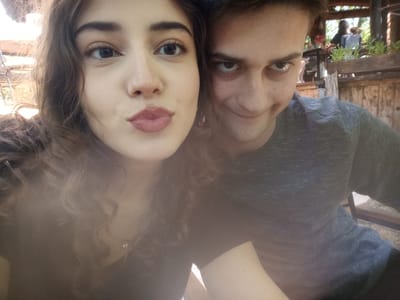 In the foreground, a young woman with dark curly hair and fair skin is captured in a close-up selfie, her lips pursed. To her right, a young man with dark hair and fair skin smiles enigmatically. In the background, out of focus, several figures are visible seated at tables under a wooden canopy, with greenery and rustic wooden structures suggesting an outdoor dining or recreational area. The overall lighting is warm and diffused, indicating it might be daytime with natural light. The location is identified as Miločani, Montenegro, implying a setting that could be a restaurant, cafe, or park within that area.