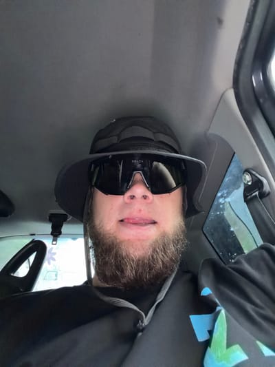A selfie is taken from the perspective of someone sitting in the driver's seat of a vehicle, likely a car. The person is wearing a black bucket hat and large, black, wraparound sunglasses with "PRADA" visible on the lens. A portion of the person's face, including their nose, mouth, and a beard, is visible. Their tongue is sticking out slightly. The interior of the car is dark, with a gray headliner and a black steering wheel visible to the left. A seatbelt buckle hangs from the ceiling. The background outside the car window on the right is blurred but suggests an outdoor environment. A hint of the exterior of the car and other vehicles are visible through the windshield.