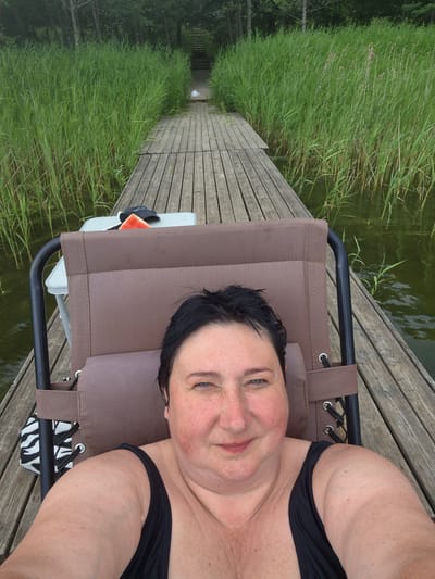 A woman is lying on a lounge chair on a wooden pier. She is wearing a black swimsuit. She is looking at the camera and smiling. The pier is surrounded by tall green grass and water. There is a small table next to her with a slice of watermelon and a black hat. In the background, there is a path leading up to stairs and trees. The scene appears to be in Rimšas, Latvia.
