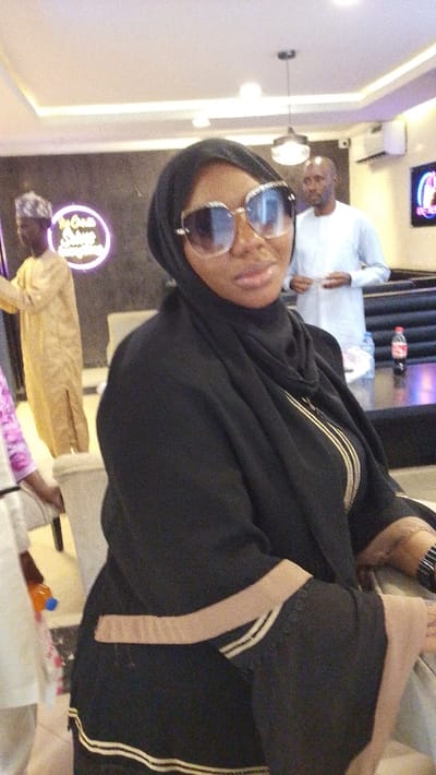 A woman wearing sunglasses and a black hijab sits in a chair. Her black garment features beige trim on the sleeves. Several individuals are visible in the background: one in a gold garment, another in white, and part of someone in pink. A neon sign is illuminated in the background, and a black table displays drinks. The room has recessed lighting and an air conditioning unit.