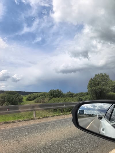 The image shows a view from inside a vehicle traveling on a road, possibly near Chernovskoe, Russia. The sky is overcast with a mix of blue and gray clouds; contrails are visible. A dark patch of precipitation is visible in the distance. A guardrail runs along the side of the road. Beyond the guardrail, a field of low-lying green vegetation is visible. The car's side mirror shows a reflection of the road behind, including other vehicles and a road sign. The reflection shows the road continuing straight. The road surface is asphalt.