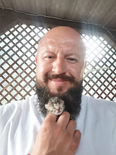 The image is a selfie taken outdoors, likely on a porch or gazebo, as indicated by the wooden lattice structure in the background. The subject is a bald, middle-aged man with a full, dark beard, wearing a white shirt. He is holding a very small, light-colored kitten in his hand, which is positioned near his mouth. The kitten appears to be sleeping or very young, with its eyes closed. The background is a blurred view of an outdoor environment through the wooden lattice. The lighting suggests it is daytime, with sunlight filtering through the gaps in the lattice. There are no other people or animals visible in the frame. The location is not discernible from the image itself.