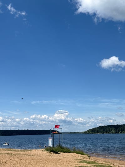 An outdoor scene at a sandy beach on a bright, sunny day. The sky is a vibrant blue with scattered white cumulus clouds. A calm, blue lake occupies the middle ground, with a dense, dark green forest lining the far shore.

In the foreground, a sandy beach slopes gently towards the water. To the right, a lifeguard stand is prominently positioned, featuring a red flag flying at the top and a sign with text and graphics on its support. Some green vegetation grows near the base of the stand.

In the lake, a person is kayaking on the left side of the frame. Further to the right, two individuals are swimming in the water. The water appears clear with small ripples on the surface.

In the sky, a small, dark object, possibly a drone or a bird, is visible in the upper left quadrant. The overall impression is one of a peaceful recreational setting at a lake.