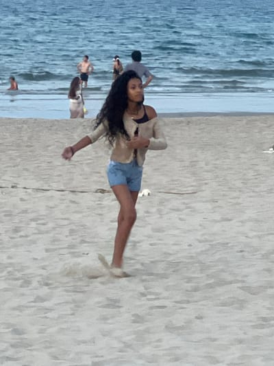This media file shows a scene at the beach with several people. The main subject is a young woman with long, dark hair, wearing a dark bikini top and light blue denim shorts. She is standing on the sand, her body oriented horizontally in the frame. She is wearing a light-colored, possibly beige, open jacket or cardigan over her bikini top and shorts. Her left arm is extended, and her hand is slightly open. She appears to be looking towards the camera or slightly past it with a neutral expression.
In the background, the ocean with gentle waves is visible on the left side of the frame. The water is a muted blue-gray, suggesting it might be overcast or late in the day. On the sand, other individuals can be seen. To the left of the main subject, a person in a gray shirt and dark shorts is partially visible. Further in the background, a person with no shirt is visible near the water's edge, and another person is walking further down the beach. The setting is clearly a sandy beach with the ocean in the background, indicating a coastal location.
The lighting suggests it might be late afternoon or early evening, as there are no harsh shadows and the overall light is soft. The mood seems relaxed, typical of a beach day. There is no visible text in the image. The scene is consistent with a beach in Da Nang, Vietnam, given the context provided.