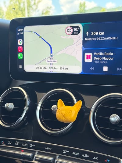 The image shows a close-up view of a car's dashboard, focusing on the infotainment screen and air vents. The infotainment screen displays a navigation interface with a map of the Egnatia Odos highway in Greece. The current time is 14:32, and the car is traveling at 107 km/h, with a posted speed limit of 130 km/h. The destination is Thessaloniki, located 209 km away, with an estimated arrival time of 20:40. The screen also shows a music player with "Vanilla Radio - Deep Flavour" playing from TuneIn. To the left of the screen, icons for a map application, music, and phone calls are visible. In front of the air vents, a bright yellow French bulldog-shaped air freshener is attached to the right vent. The lower part of the image shows car climate control buttons, including "AUTO", "MENU", "REAR", "SYNC", and "A/C". The environment appears to be outdoors, with sunlight filtering through trees in the background, suggesting a daytime shot. The geographical location is identified as the Egnatia Odos highway, which passes through the Pamvotida Municipal Unit in Greece.