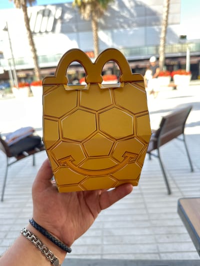 An outdoor shot shows a person's hand holding a yellow McDonald's Happy Meal box that features a hexagonal pattern and two arching handles. The background is blurred but suggests a street scene with palm trees and buildings, possibly in Platja d'Aro, Spain. The box is positioned in the center of the frame, obscuring the rest of the person's body.