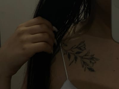 This scene, reported to be in Vakhitovo, Russia, presents a close-up, oblique view of a person's upper torso, focusing on the right shoulder and adjacent neck area. The individual exhibits a light complexion and has long, dark hair. A left hand, featuring long, light-colored fingernails, is visible on the upper left, appearing to grasp or adjust strands of the dark hair near the neck. On the right clavicle/shoulder, a black-ink tattoo depicts a botanical design, featuring a central branch with multiple leaves and two distinct flower blossoms. A thin white spaghetti strap from a garment, consistent with a camisole or tank top, crosses the lower right of the frame, partially obscuring the lower portion of the tattoo. The background is a uniformly light, indistinct surface, indicative of an indoor setting with low ambient illumination.