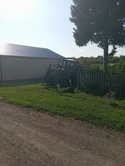 The image depicts a rural scene in Novo-Kopylovo, Russia, on a bright, sunny day. In the foreground, a gravel road runs diagonally across the bottom of the frame. To the left of the road is a patch of vibrant green grass.
Dominating the mid-ground is a green tractor with a front-end loader. It is positioned on the grass, partially obscured by a wooden fence. The fence, made of vertical wooden planks, runs along the right side of the image and extends into the background. Behind the tractor and fence, lush green foliage and trees are visible, indicating a natural environment.
To the left of the tractor and fence, a large, modern building stands. It features a light-colored, horizontal siding and a dark, corrugated metal roof. A large, mature tree with dense foliage is situated behind the tractor and to the right of the building, casting a shadow. The sky above is clear and a pale blue. No people are present in the image.
