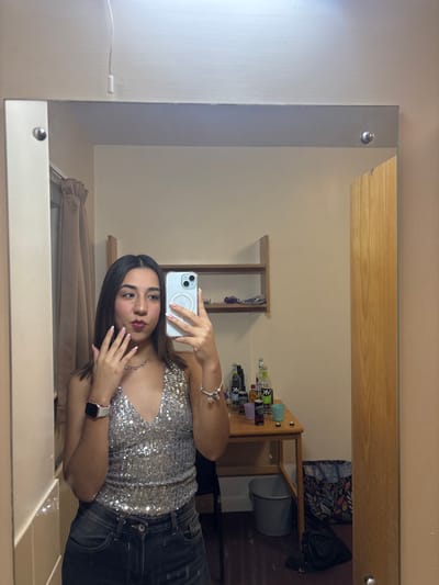 A mirror selfie capturing a young woman. She is dressed in a silver sequined top and dark jeans. Her phone is visible in her hand as she takes the photo. The reflection shows a tidy room with a desk holding various items, including what appears to be alcohol bottles. There's also a shelf and bags in the background. The scene is well-lit, highlighting the sparkle of her top and the organized space behind her.