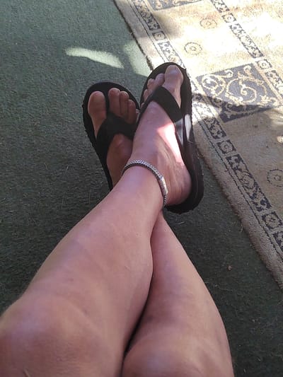 The image shows a close-up view of a person's legs and feet, wearing black flip-flops and a silver bracelet. The legs are crossed, with the feet resting on a green surface, possibly artificial grass, with a patterned rug partially visible in the background. Sunlight creates a shadow on the green surface. The scene is outdoors, with the texture of the rug suggesting it is placed on a patio or balcony. The image does not contain enough information to determine the city of Yuzhnyy, Russia.