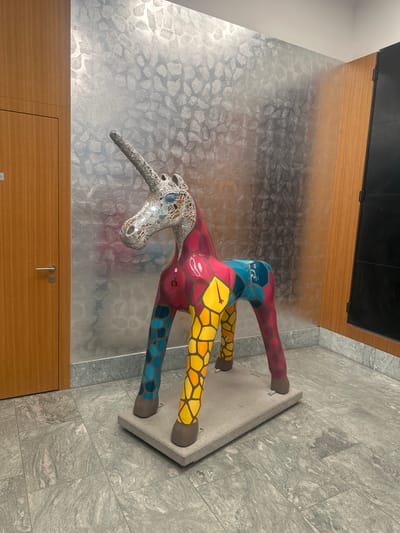 The image depicts a mosaic unicorn sculpture standing on a concrete base indoors. The unicorn's head is covered in a mosaic of small mirror-like tiles, while its body and legs are painted in geometric patterns of red, yellow, blue, and green. The sculpture is positioned against a wall with a textured, metallic-looking silver covering. In the foreground, there is a light brown wooden structure, possibly a cabinet or paneling. The floor is tiled in a light gray stone pattern. The sculpture is located in the city of Dübendorf, Switzerland.