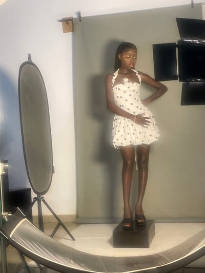 In a studio setting, a young Black woman stands posing on a dark rectangular platform. She is wearing a white polka-dot halter dress that ends mid-thigh, and black platform heels. She has on a necklace and bracelets. Her eyes are closed, and her head is tilted downwards, with her lips slightly parted. The background consists of a grey backdrop and studio lighting equipment, including a large oval reflector on a tripod to her left and two rectangular lights to her right. The floor appears to be tiled. The studio is located in South Ropp, Nigeria.