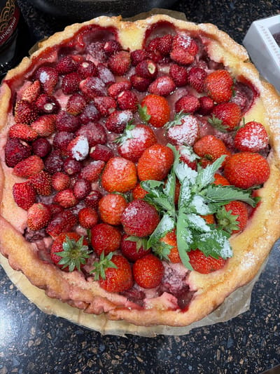 A high-angle, close-up shot captures a rustic strawberry tart, likely homemade, presented on a dark, speckled countertop. The tart is generously filled with a mix of ripe strawberries, ranging in size from small wild strawberries to larger cultivated ones. Some of the berries are dusted with powdered sugar, giving them a frosted appearance. A few sprigs of fresh mint, also lightly coated in sugar, are placed in the center as a garnish. The crust of the tart is golden brown and appears slightly uneven, suggesting a homemade quality. The filling is a vibrant red, indicating the presence of strawberry jam or cooked strawberries.
In the upper left corner, a partially visible white box with a decorative pattern is present, hinting at a possible purchase or presentation of baked goods. The overall scene is indoor, with the dark countertop providing a strong contrast to the bright colors of the fruit and crust. The lighting is natural, creating soft shadows and highlighting the textures of the tart. There are no people visible in the frame. The image is devoid of any specific time of day or weather cues. No other discernible location cues are present beyond the countertop. The image evokes a sense of homemade comfort and the freshness of summer fruits. There is partially visible text on a dark label in the bottom left corner, but it is too indistinct to be read. The provided location context of "Северка, Russia" does not offer specific visual information within the image.