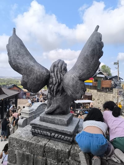 The image shows a large stone statue of a winged figure, possibly an angel or a mythological creature, in a public space with a festival or market atmosphere. The statue is dark gray and weathered, with large wings outstretched and a carved, detailed head. The statue is positioned on a stone pedestal, which is part of a larger stone structure. In the foreground, three young individuals are partially visible, two girls are crouching and looking at something off-camera, and a third person's legs are visible from the knees down.

Behind the statue, there is a lively scene of a town or village. Several wooden stalls and buildings with traditional architecture are visible, suggesting a historical or themed setting. Some people are walking around and interacting, some are seated at tables. The stalls have signs that suggest food and drink vendors, one with text that might be "Rancho da Capelheira". The overall environment appears to be outdoors, with a bright, partly cloudy sky overhead and a landscape with trees and buildings visible in the distance. The architecture and setting strongly suggest a historical reenactment or a themed village, potentially at a festival. The presence of the statue and the themed stalls indicates this location is within Óbidos, Portugal, known for its medieval castle and historic town center, which often hosts events and festivals.