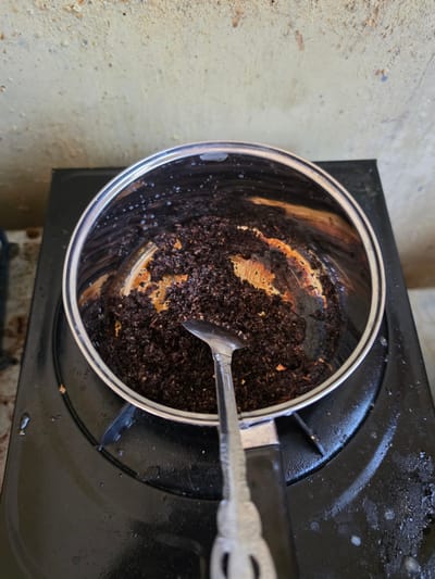 The image shows a close-up, top-down view of a burnt pot on a black gas stove. The pot, made of stainless steel, is filled with dark brown, granular residue, likely burnt coffee grounds. The sides of the pot show significant signs of burning, with areas of orange and black charring. A silver spoon rests inside the pot, also covered in the residue. The stove top appears greasy and stained. In the background, a textured, yellowish wall with some blue and rust-colored splotches is visible. The setting appears to be an outdoor or semi-outdoor kitchen area, possibly in Pintomakmur, Indonesia, given the context.