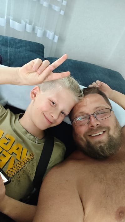 A close-up, selfie-style photo shows two males lying down, likely on a bed or sofa, with a portion of a dark blue cushion and white curtains in the background. The person on the left is a young boy with short, blonde hair, wearing a green t-shirt with yellow lettering and a cross necklace. He is smiling and making a peace sign with his right hand, which is positioned near his forehead. The person on the right is an adult male with a beard and glasses, also smiling. He is wearing glasses and appears to be shirtless. The lighting suggests it is daytime, possibly indoors.